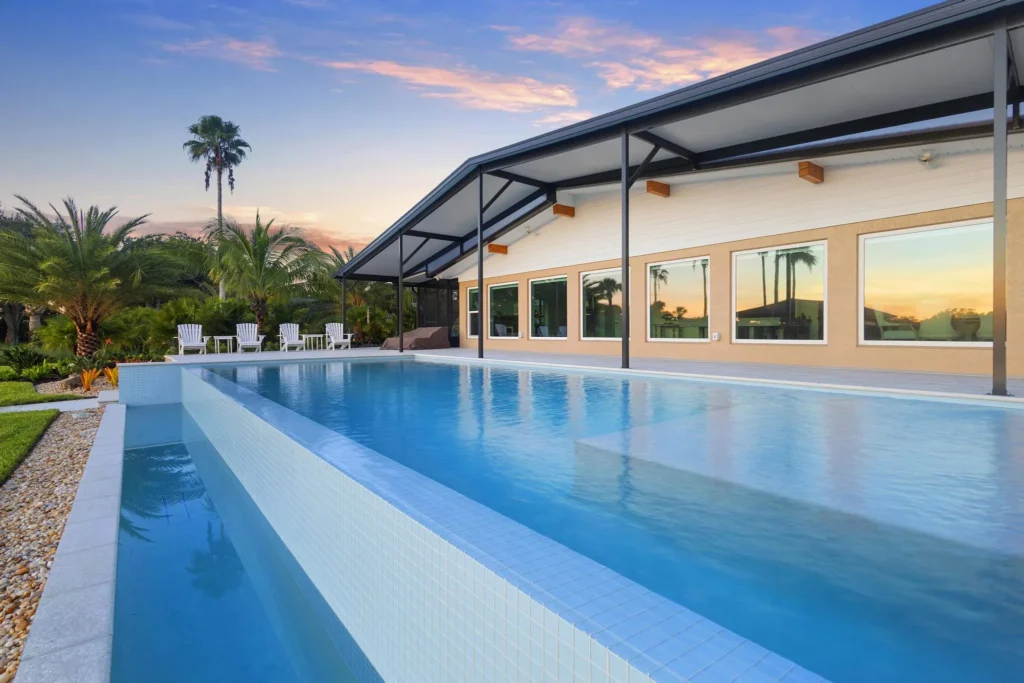 An elevated lap pool with a glass-tile vanishing edge runs alongside a modern home at dusk with palm trees in the background.