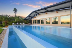 An elevated lap pool with a glass-tile vanishing edge runs alongside a modern home at dusk with palm trees in the background.