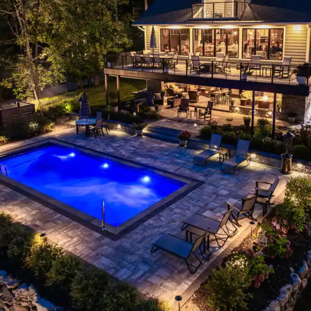 A rectangular backyard pool glowing blue at night beside a stone patio and multi-level deck.