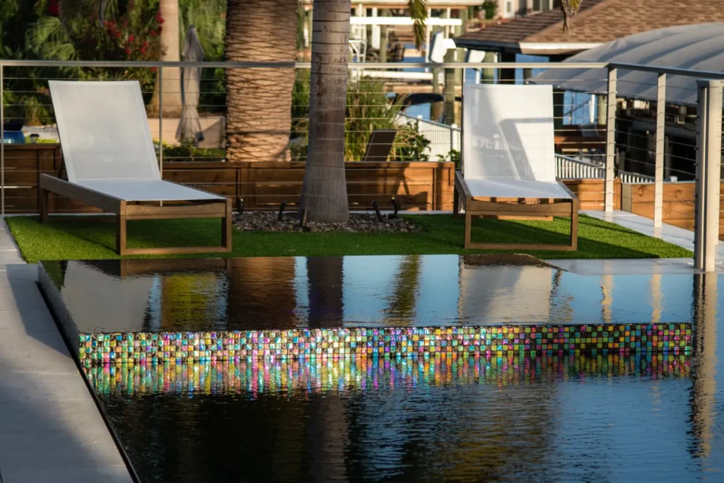 A vanishing-edge pool wall with vibrant mosaic tile reflecting in the water beside a canal.