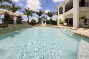 A large rectangular backyard pool with a raised spa and multiple wall-mounted water scuppers, framed by palm trees and a two-story white house.