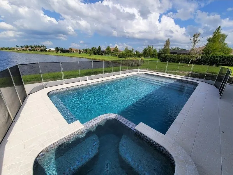 A rectangular pool with an attached spa overlooks a lakefront backyard, enclosed by a removable safety fence.