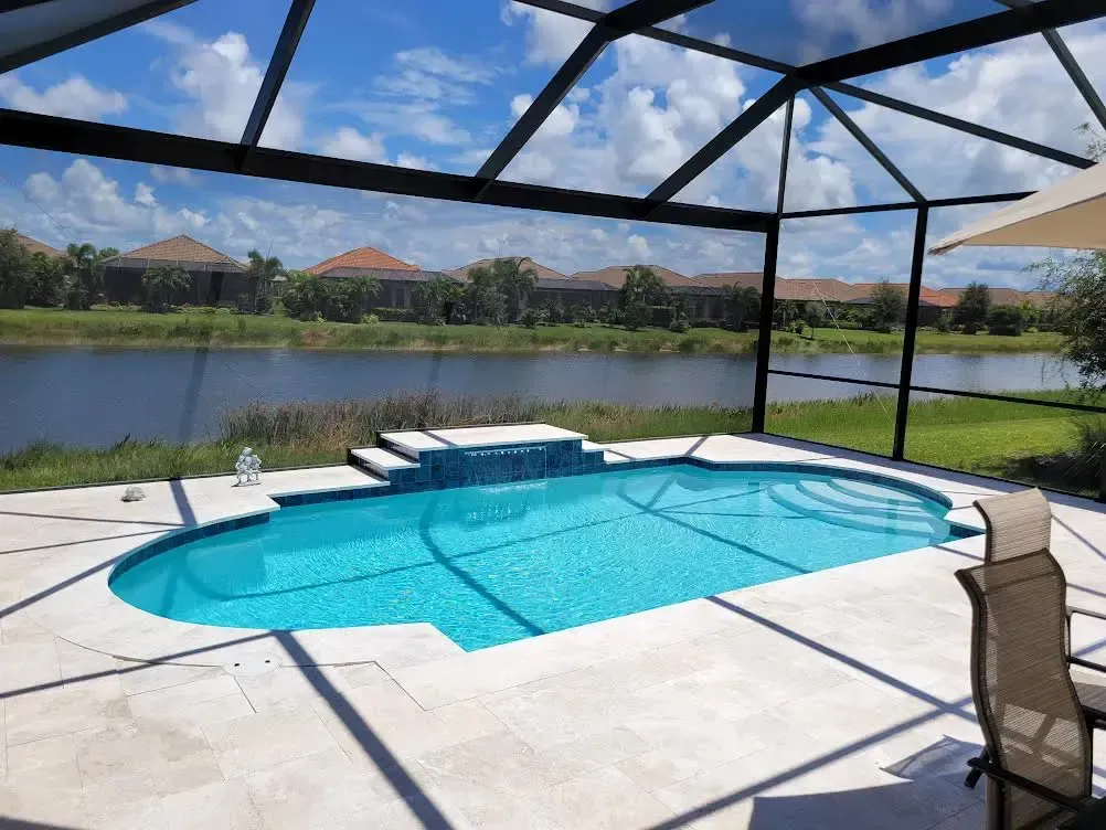 A freeform pool with a small raised water feature sits on a travertine deck under a screened enclosure overlooking a canal.