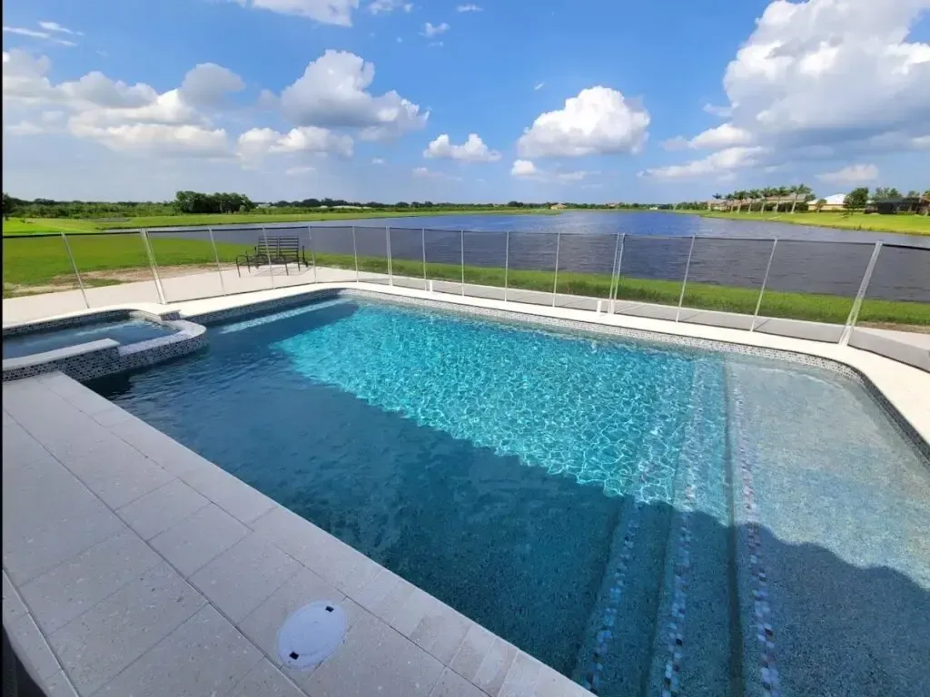 An L-shaped pool with an attached raised spa overlooks a lake under a pool safety fence.