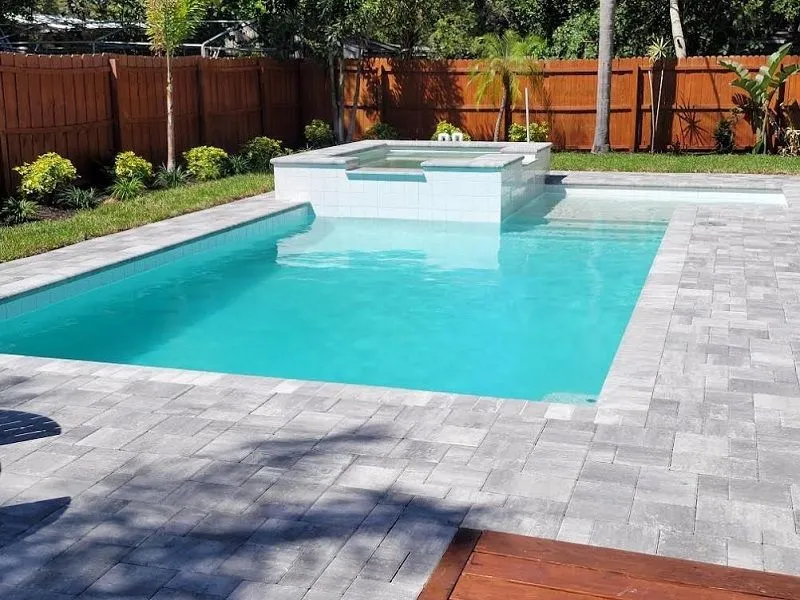 A rectangular backyard pool with a raised tiled spa at one end, surrounded by gray paver decking and a wood privacy fence.