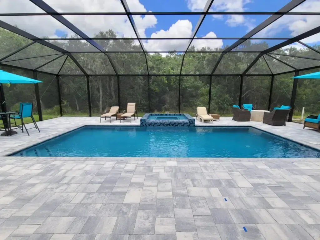 A rectangular pool with a raised square spa under a screened lanai, surrounded by a paver patio and wooded backyard.