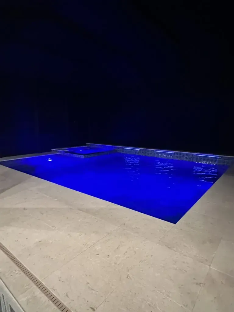 A rectangular backyard pool lit blue at night with a raised spa and small water scuppers along the far wall.