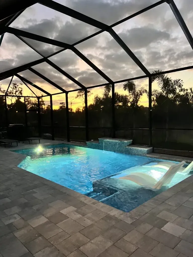 A screened pool with a raised spa spilling into the water at dusk, an in-pool lounger visible near the steps.