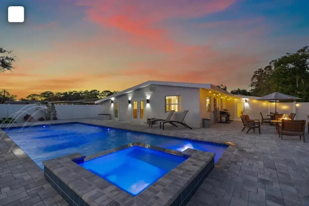 A rectangular pool with an attached raised spa glowing blue at dusk, set on a paver patio behind a single-story home.