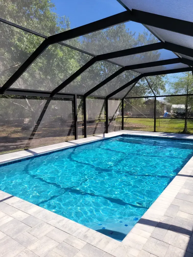 A rectangular pool with turquoise water under a screened patio enclosure, surrounded by pavers and trees.