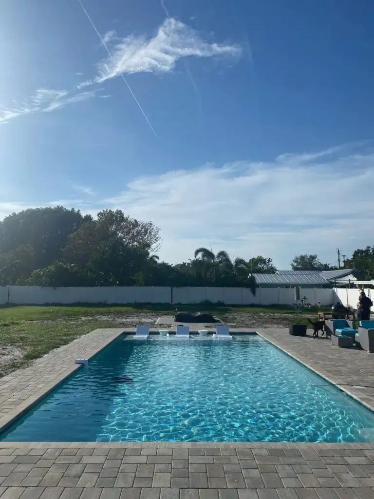 A rectangular backyard pool with submerged loungers and a paver patio under a sunny sky, people visible in the distance.