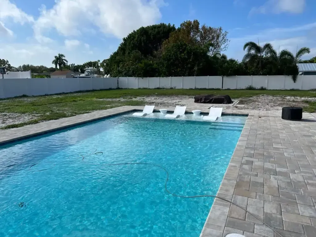 A rectangular backyard pool with built-in white loungers and a paver deck, yard still being landscaped.
