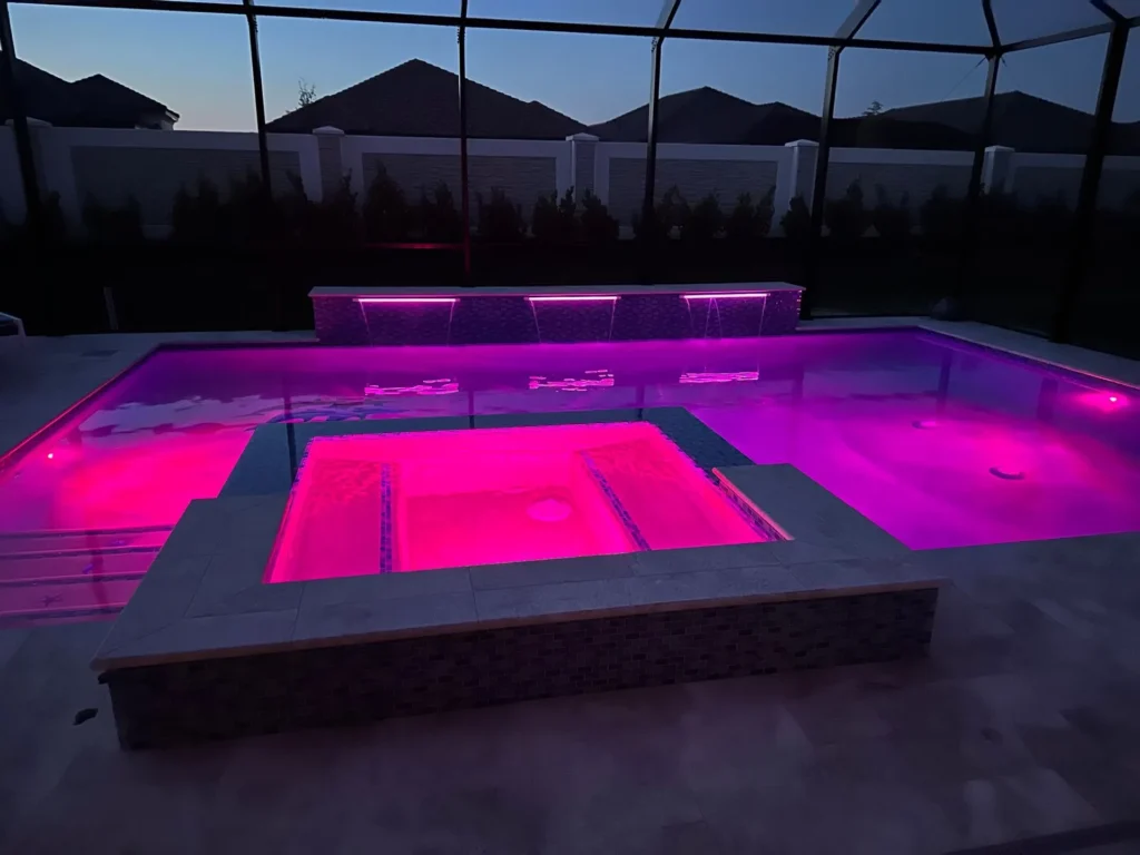 A raised spa glowing pink with LED lights sits in front of a pool with a scupper waterfall, under a screened enclosure at dusk.