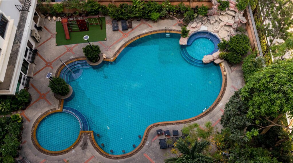 Aerial view of a curvy resort-style pool complex with a small round spa, waterfall rocks, and a playground beside the deck.