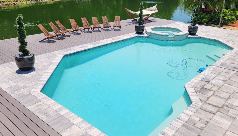 A geometric pool with a raised corner spa and paver deck sits beside a canal lined with wooden loungers and a hammock.