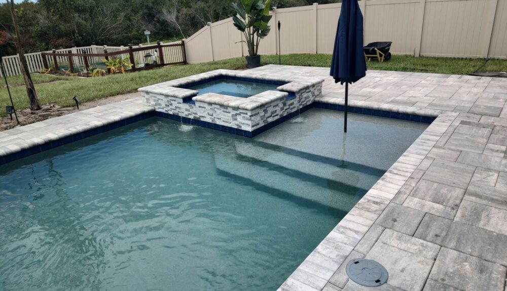 A rectangular backyard pool with a raised stacked-stone spa spilling into a tanning ledge, set on paver decking with a privacy fence behind.