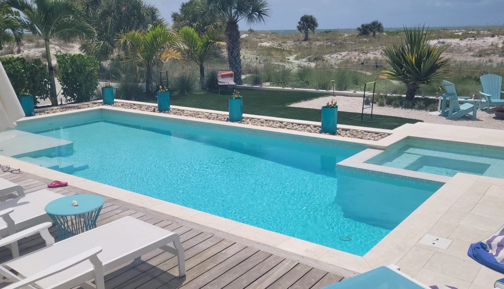 A rectangular pool with an attached spa overlooks a beach dune landscape with palm trees.