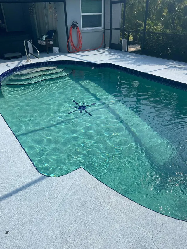 A small freeform pool with a tiled waterline under a screened lanai, with an automatic pool cleaner floating in the water.