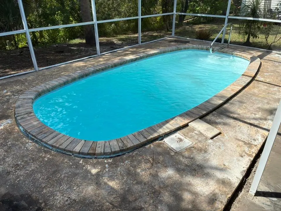 An oval fiberglass pool with brick coping under a screened enclosure, surrounded by worn concrete decking.
