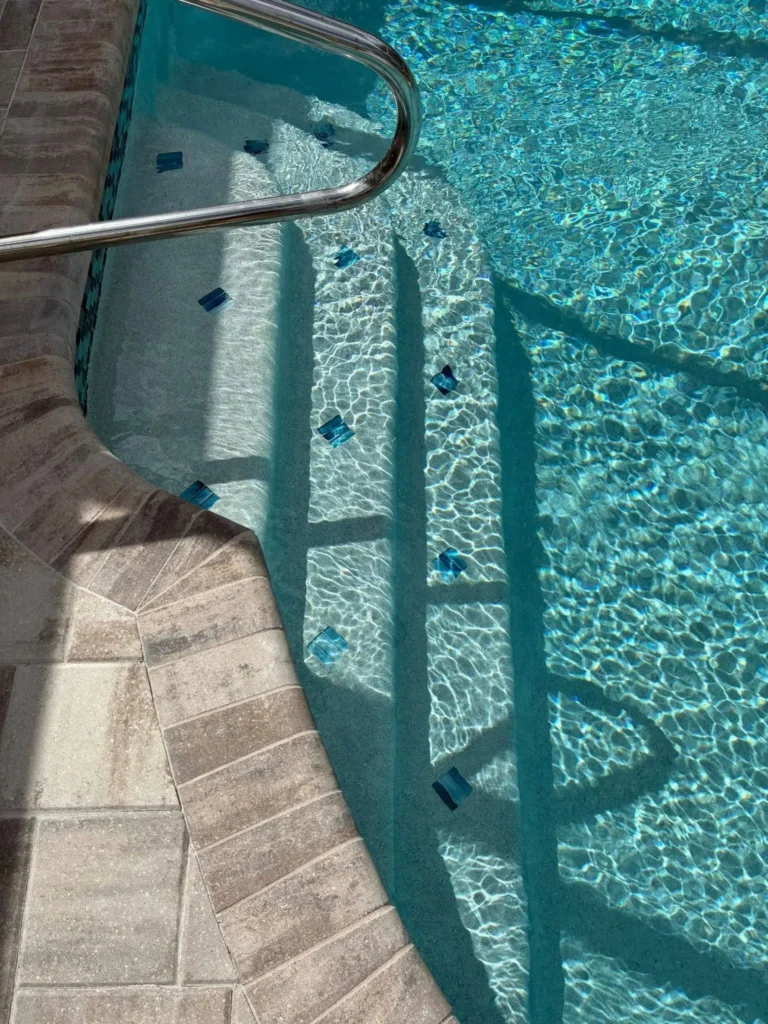 Close-up of pool entry steps with a stainless handrail and small decorative tile accents underwater.