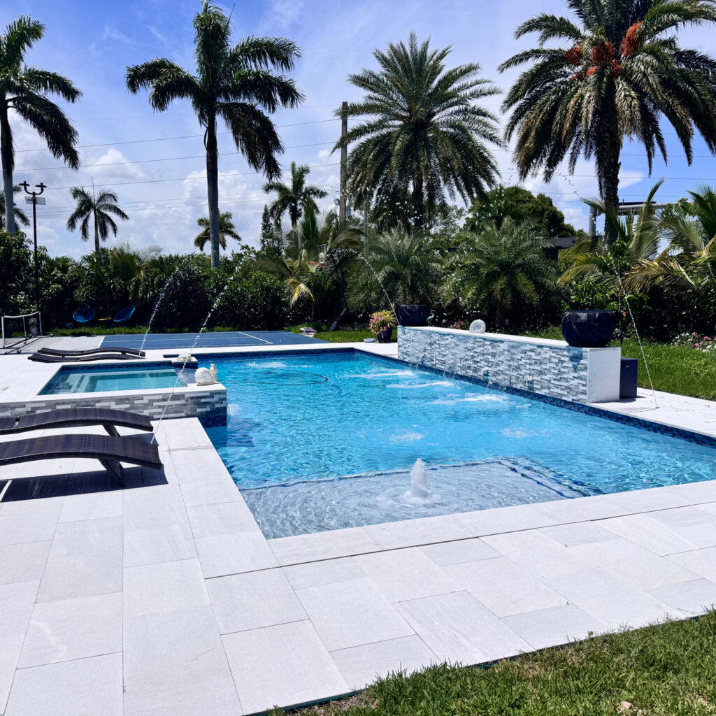 A rectangular backyard pool with a raised spa, tiled accent wall, and dancing water jets surrounded by palm trees.