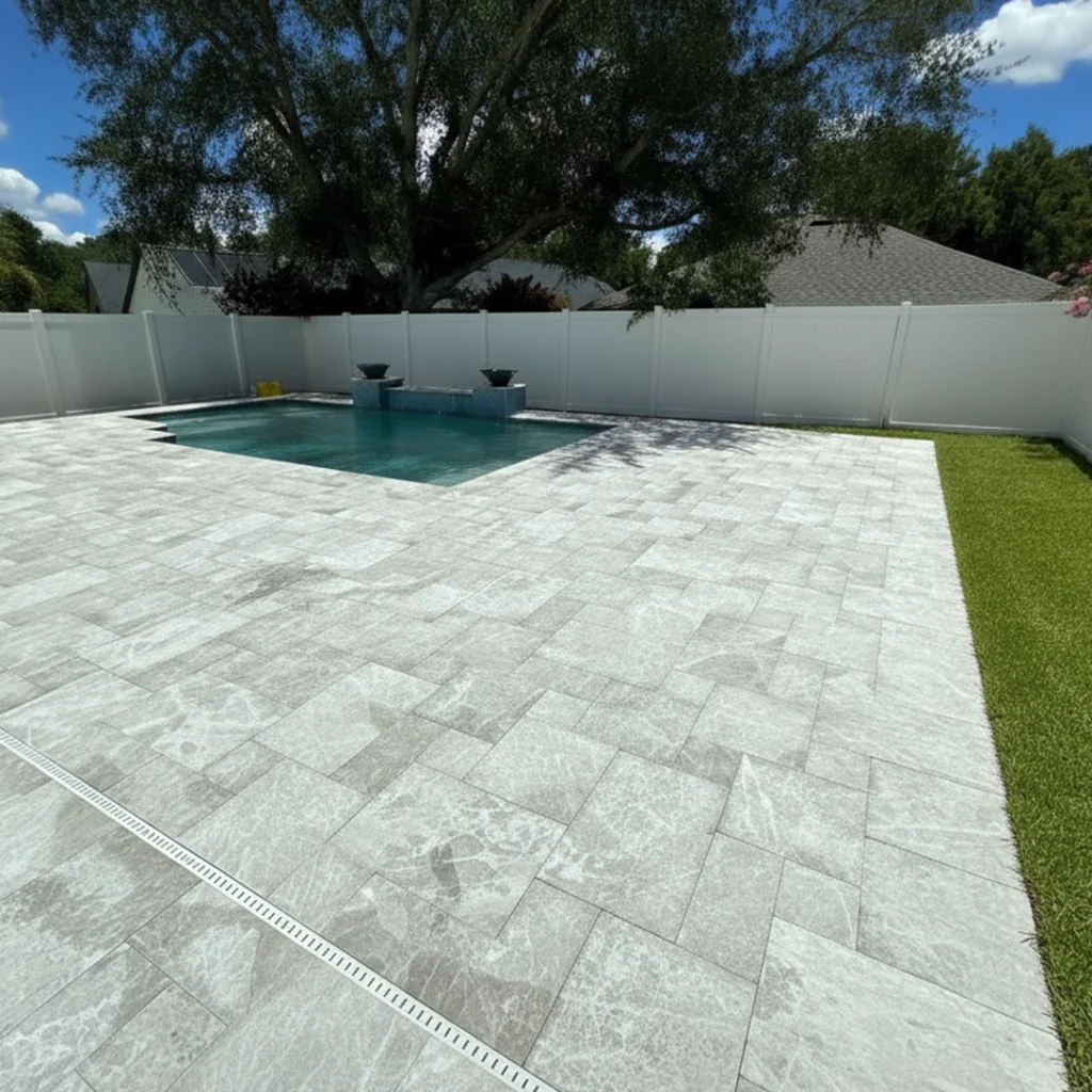 A small rectangular pool with two decorative spillway bowls surrounded by a large gray paver patio and turf strip in a fenced backyard.