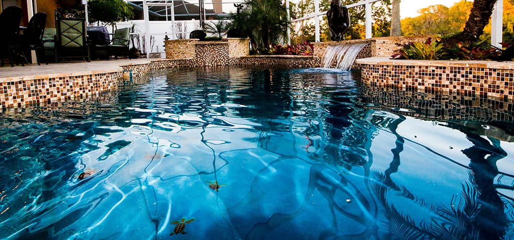 A dark-finished freeform pool with a tiled raised wall and small waterfall under a screened enclosure at dusk.