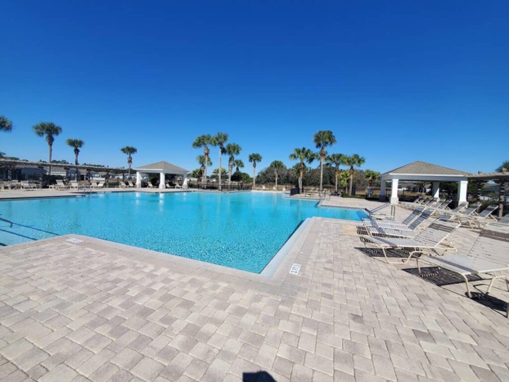 A large rectangular community pool with turquoise water surrounded by paver decking, lounge chairs, palm trees, and gazebos under a clear blue sky.