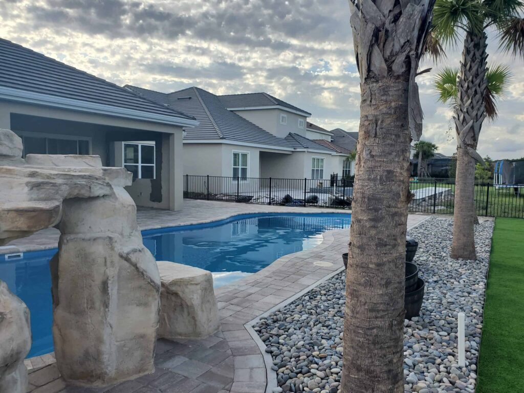 A small freeform pool with a large rock waterfall feature beside a paver patio and palm trees in a suburban backyard.