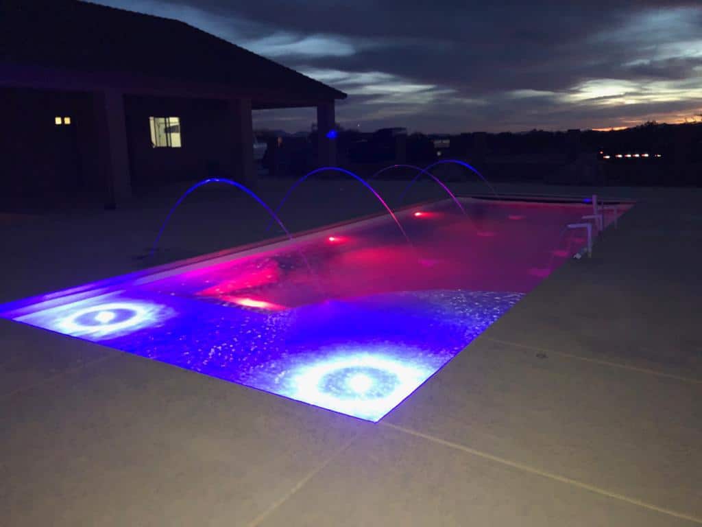 A rectangular backyard pool lit with vivid blue and red LED lights and arching water jets at dusk.