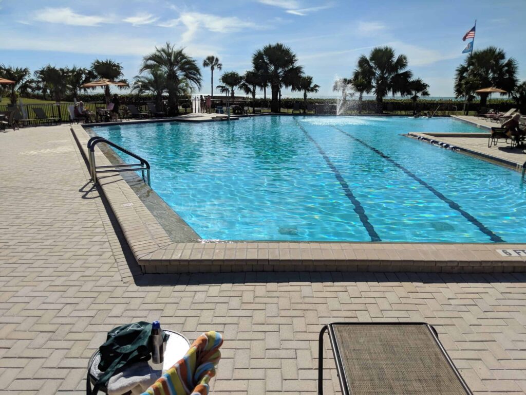 A large community lap pool with lane markers and a fountain, surrounded by palm trees and paver decking.