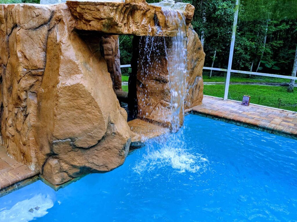 A large artificial rock waterfall cascades into the corner of a pool under a screened enclosure.