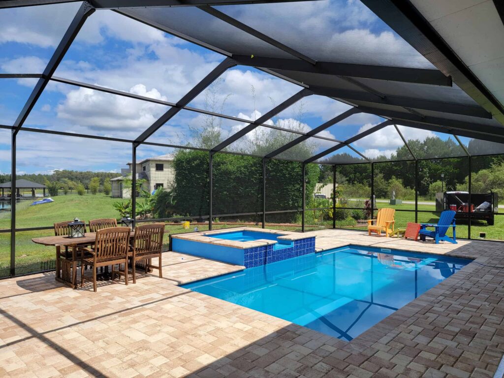 A rectangular pool with a raised spa under a screened patio enclosure with paver decking and outdoor dining set.