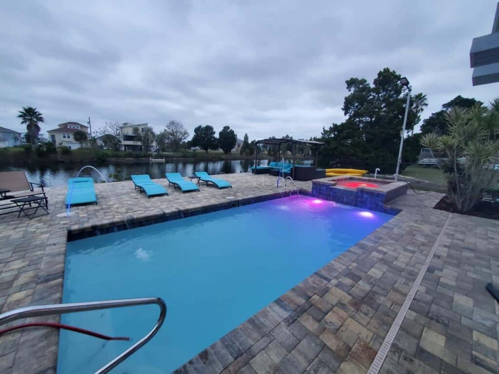 A rectangular pool with a raised spa lit purple and red at dusk beside a canal.