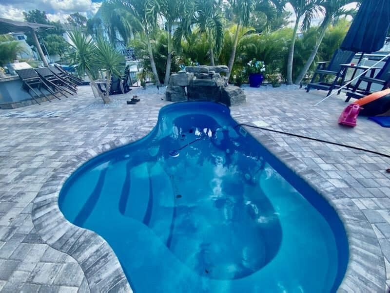 A freeform fiberglass pool with a rock waterfall feature surrounded by a paver patio and tropical palms.