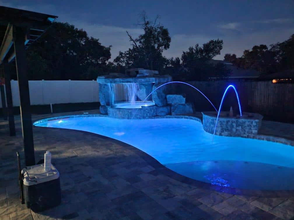 A freeform pool at night lit blue with two arching water jets and a raised stone spa with a waterfall.