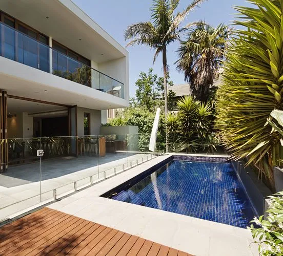 A narrow lap pool with dark blue glass tile runs alongside a modern multi-story home with a glass balcony.