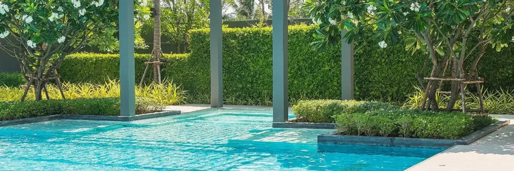 A resort-style pool framed by pergola columns and lush planted islands with a tall hedge backdrop.
