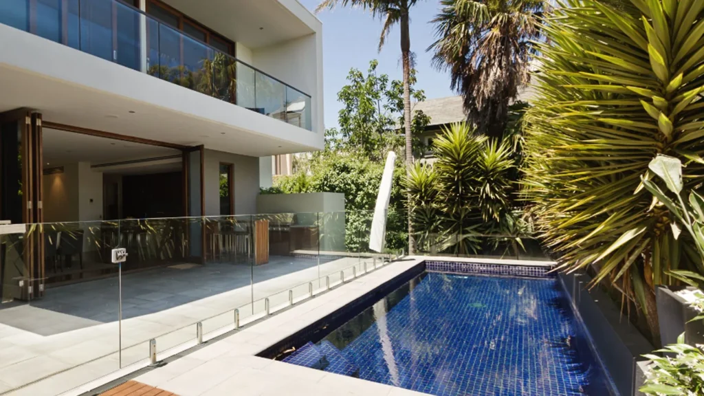 A narrow blue-tiled lap pool runs along a modern two-story home with a glass safety fence and tropical landscaping.