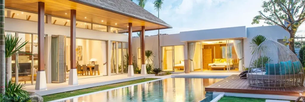 A modern villa at dusk with a narrow lap pool running alongside a covered terrace and wooden deck.