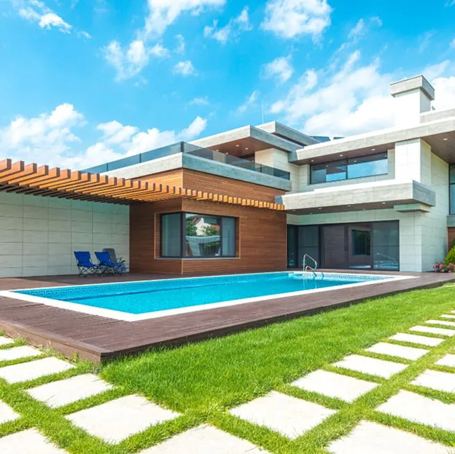 A modern house with wood-slat pergola overlooks a small rectangular pool on a wood deck beside a manicured lawn.