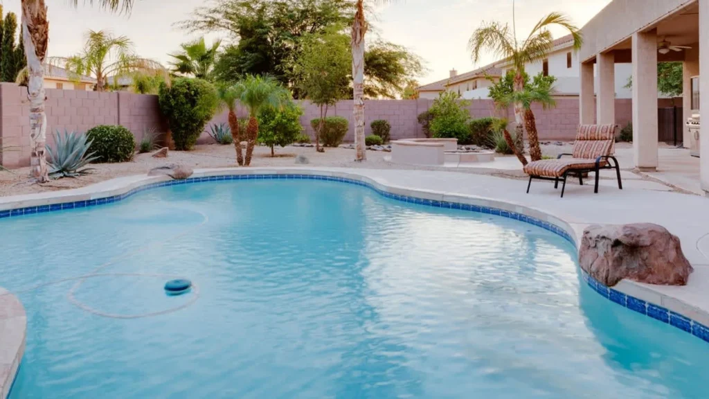 A freeform backyard swimming pool with a rock accent at dusk, surrounded by desert landscaping and a block wall.