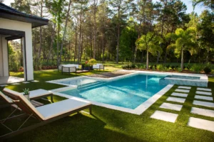 A rectangular backyard pool with a raised spillover spa, surrounded by artificial turf, pavers, and lounge chairs with trees in the background.