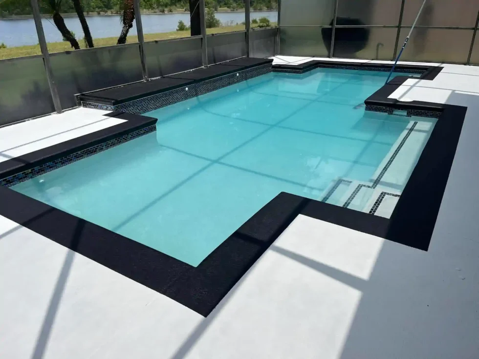 An L-shaped pool with dark coping and a tanning ledge sits under a screened enclosure overlooking a lake.