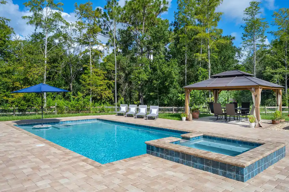 A rectangular backyard pool with an attached raised tiled spa, surrounded by a paver patio, loungers, umbrella, and a wooden gazebo with dining set, backed by trees.