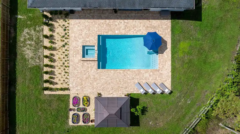 Aerial view of a small rectangular backyard pool with an attached spa and blue umbrella on a paver patio.