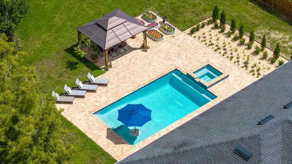 Aerial view of a rectangular backyard pool with an attached raised spa, paver deck, gazebo, and loungers.