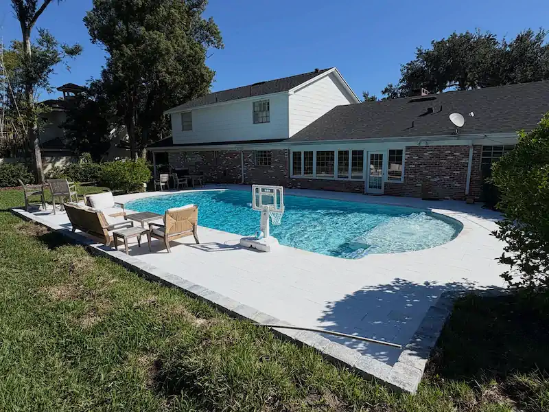A freeform backyard pool with white paver decking, patio furniture, and a basketball hoop behind a two-story home.