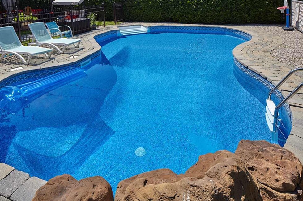 A freeform vinyl-liner pool with a paver patio, loungers, and decorative boulders in a suburban backyard.