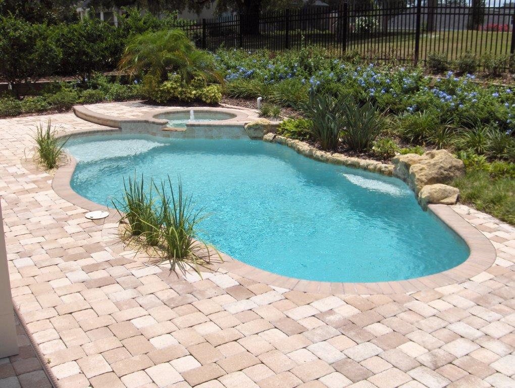 A freeform backyard pool with a raised spa and fountain jet surrounded by paver decking and landscaping.